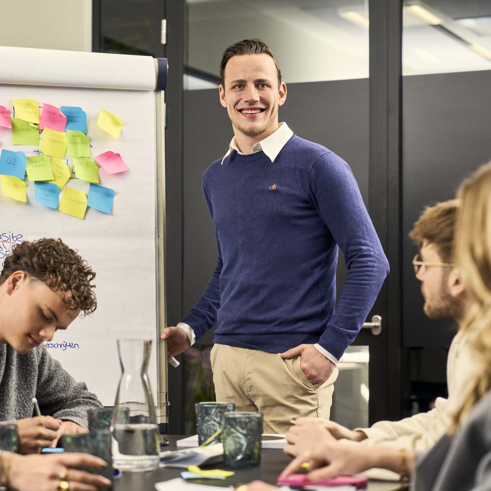 Man kijkt blij naar de camera. Hij staat langs een aantekeningenbord met post-its